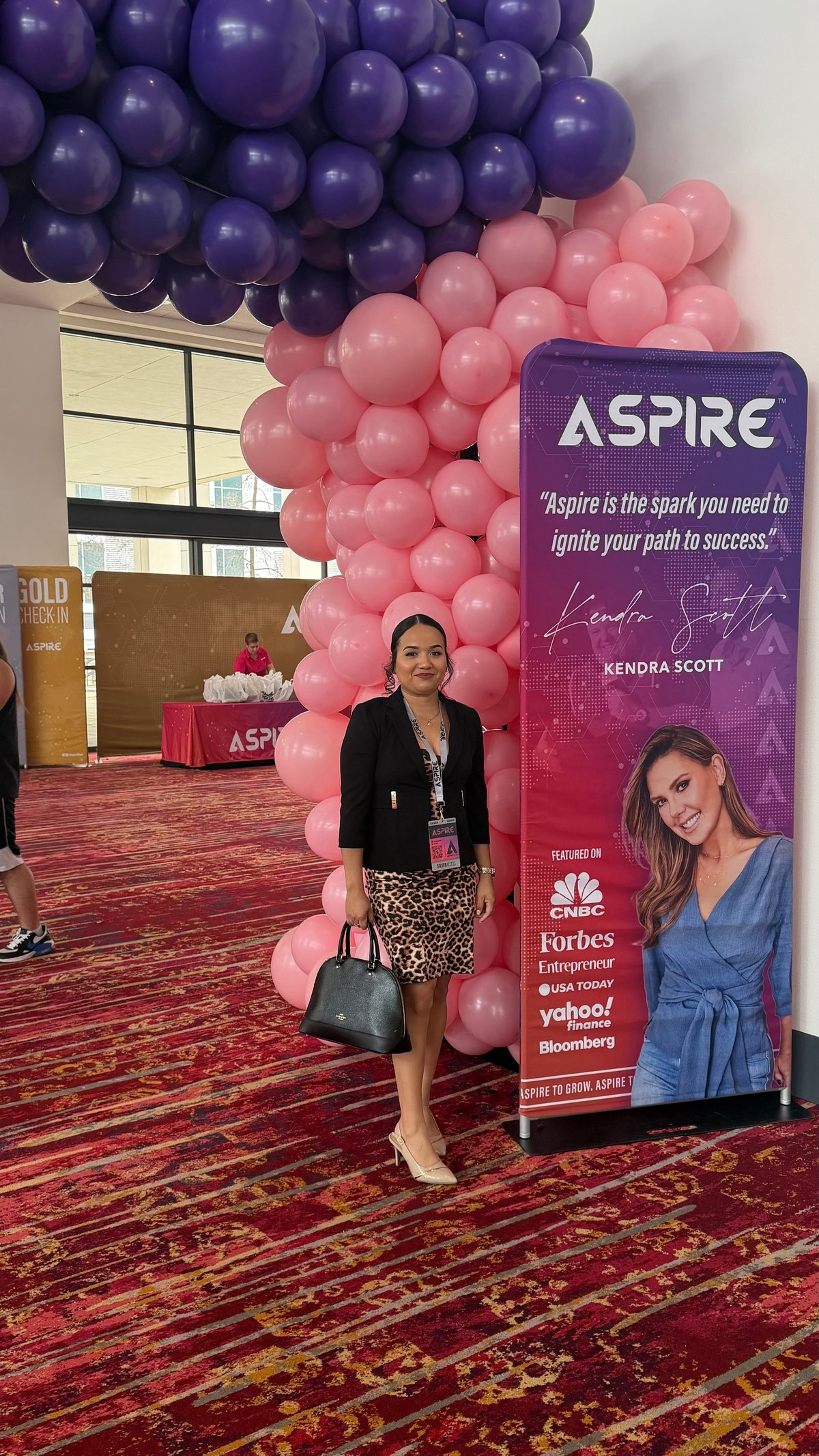 Woman posing at an event with a large ASPIRE banner and purple and pink balloon backdrop behind her